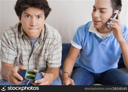 Close-up of a young man playing video game with another young man talking on a mobile phone beside him