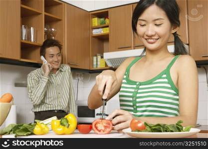 Close-up of a young woman cutting vegetables in the kitchen with a young man talking on a mobile phone