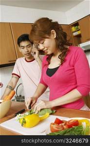 Close-up of a young woman cutting vegetables with a mid adult man talking on a mobile phone beside her