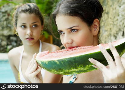 Close-up of a young woman eating a slice of watermelon with another young woman looking at her