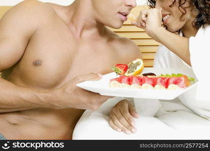 Close-up of a young woman reclining on the bed with a young woman and holding a tray of fruit salad