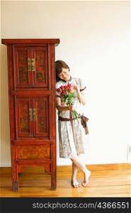 Close-up of a young woman standing near a cabinet and holding a bouquet of red roses