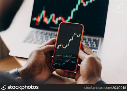 Close up of african man stock trader holding mobile phone against laptop monitor, investor analyzing indexes compares financial charts and trading online investment data on cryptocurrency stock market. African man stock trader holding smartphone with financial charts and trading online investment data