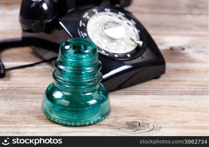Close up of an antique electric glass insulator with vintage manual dial phone in background. Rustic wood underneath telecommunication objects.