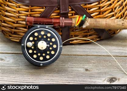 Close up of an antique fly fishing reel, rod, and artificial flies in front of creel with rustic wood underneath. Layout in horizontal format.