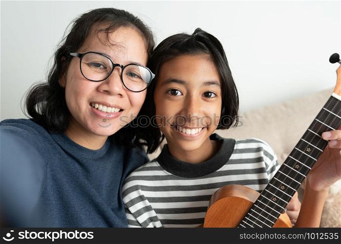 Close-up of an Asian mother and daughter taking a selfie and smiling while sitting on the sofa. The little girl holding a ukulele with her left hand in the living room at her home.