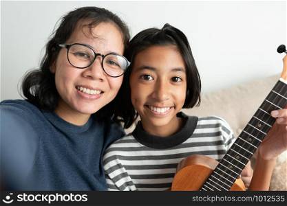 Close-up of an Asian mother and daughter taking a selfie and smiling while sitting on the sofa. The little girl holding a ukulele with her left hand in the living room at her home.