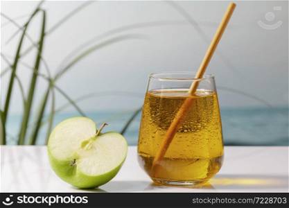 Close up of an enticing cold glass of apple juice with a straw next to half apple on a table on an out of focus background. Holiday and vegan food concept.. an enticing cold glass of apple juice with a straw next to half apple on a table. vegan food concept.