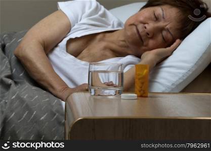 Close up of an open bottle of medicine and a glass of water on nightstand with senior woman sleeping in background. Sickness concept.