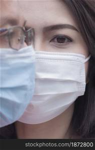 Close up of Asian couple faces in medical face masks, focus at woman?s eye looking at camera with blurred side of man?s face on foreground