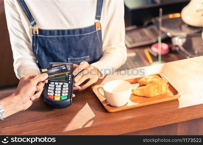 Close-up of asian customer using his credit card with contactless nfs technology to pay a barista for his coffee purchase at a cafe bar.