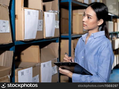 Close up of Asian smart businesswoman working with product inventory and writing on clipboard in distribution warehouse. For business warehouse inventory and logistic concept.