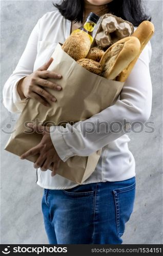 Close up of asian smile woman housewife hold grocery bag with egg cooking oil and variety of bread on grey vintage loft background. Bakery grocery and domestic life lifestyle concept for delivery.