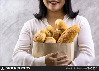 Close up of asian smile woman housewife holding variety bread in disposable paper bag on grey vintage loft background. Bakery food and drink grocery and domestic life lifestyle concept for delivery.