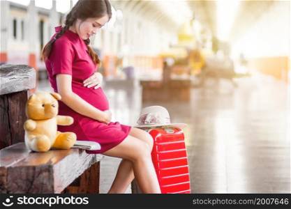 Close-up of Asian woman pregnant red dress sitting on a bench at carrying red luggage and Teddy Bear at railway station travel,traveler with backpack in summer Holiday concept Thailand