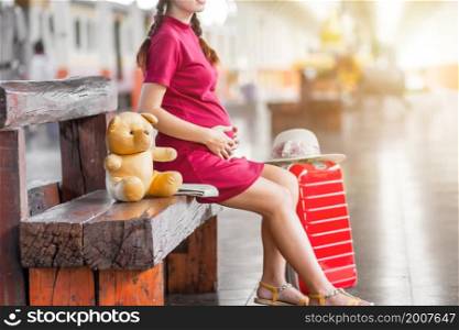 Close-up of Asian woman pregnant red dress sitting on a bench at carrying red luggage and Teddy Bear at railway station travel,traveler with backpack in summer Holiday concept Thailand