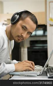 Close-up of Asian young adult man wearing headphones working on laptop and looking at viewer.