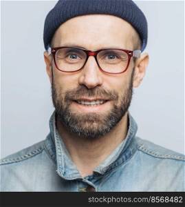 Close up of attractive blue eyed unshaven male witth pleasant appearance has delighted look, wears denim jacket, glasses and hat, isolated over white background, expresses positive emotions.