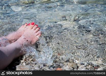 Close up of attractive woman feet with red nail polish in the crystal clear water, beach coastline