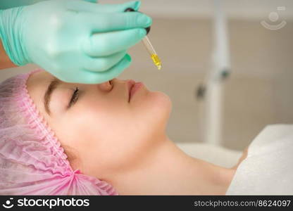 Close up of beautician holding pipette and applying oil serum drop on the face of a young woman in spa salon. Close up of beautician holding pipette and applying oil serum drop on the face of a young woman in spa salon.