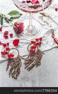 Close up of beautiful earrings on light table with red currants, rose flower and ch&agne glass. Jewelry and drink concept. Top view.