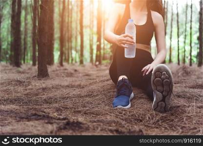 Close up of beauty sport woman resting and holding drinking water bottle and relaxing in middle of forest after tired from jogging. Girl sitting and looking attraction view. Workout lifestyle concept