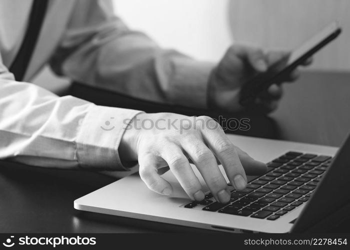 close up of businessman working with mobile phone and laptop computer on wooden desk in modern office,black and white