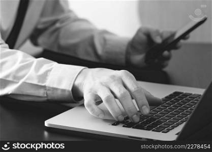 close up of businessman working with mobile phone and laptop computer on wooden desk in modern office,black and white