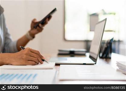 Close up of businesswoman is searching data on smartphone while working on laptop and writing notes.