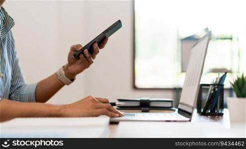 Close up of businesswoman searching business data on smartphone while working on laptop in office.