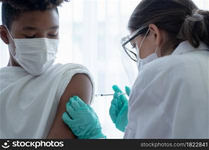 Close up of Caucasian woman doctor wear mask, face shield and gloves is giving injections or vaccines to arm of African American boy at hospital. Preventing spread of COVID-19 by vaccinating people