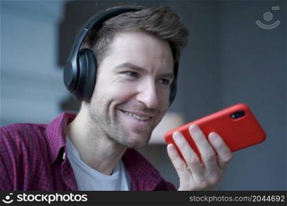 Close up of cheerful young german man in wireless headphones holding smartphone in hand with microphone close to his face during mobile conversation with friend, recording audio message. Cheerful young german man in wireless headphones holding smartphone in hand, recording audio message
