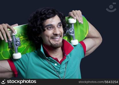 Close-up of cheerful young man holding skateboard behind head against black background