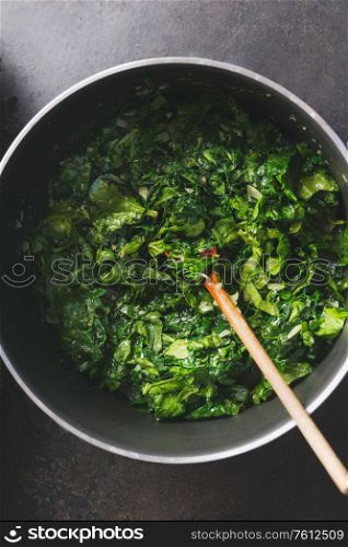 Close up of chopped stewed spinach in cooking pan with wooden spoon on rustic kitchen table. Top view. Vegetarian healthy eating concept.