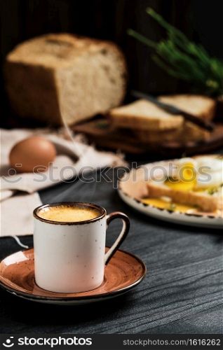 Close-up of cup of espresso coffee with rising steam on black wooden table. On blurred background, soft-boiled egg (poached) in slice of bread, with butter cream and herbs. Breakfast idea