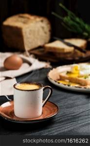 Close-up of cup of espresso coffee with rising steam on black wooden table. On blurred background, soft-boiled egg (poached) in slice of bread, with butter cream and herbs. Breakfast idea