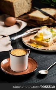 Close-up of cup of espresso coffee with rising steam on black wooden table. On blurred background, soft-boiled egg  poached  in slice of bread, with butter cream and herbs. Breakfast idea