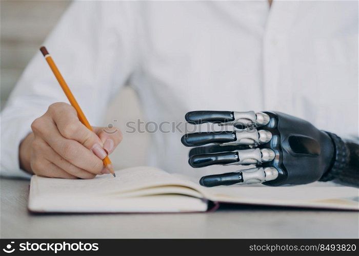 Close up of cyber arm and healthy limb on the notebook. Handicapped young woman with bionic prosthesis is studying remotely. Disabled girl is writing a book with pencil at the desk. Amputee lifestyle.. Cyber arm and healthy limb on the notebook. Woman with bionic prosthesis is studying remotely.