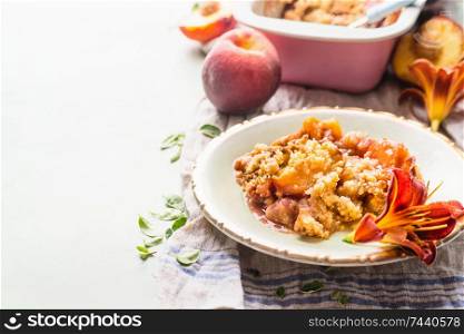 Close up of delicious peach cobbler dessert in plate on light background with baking pan and fresh peaches . Copy space