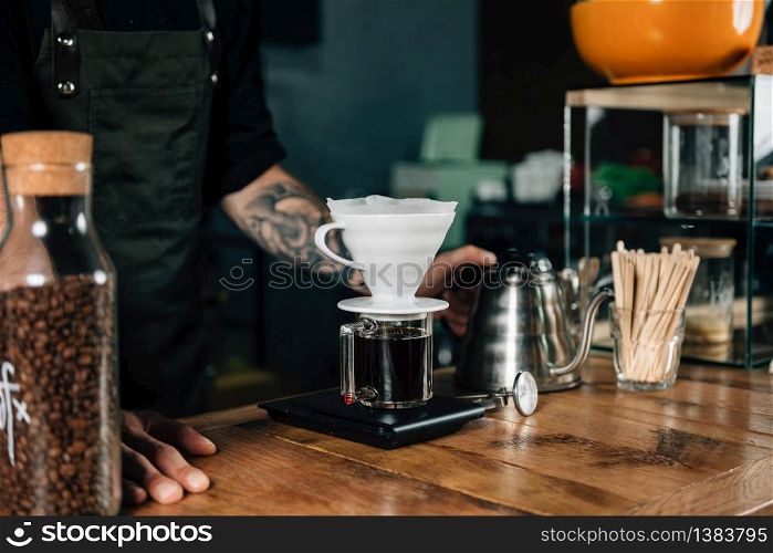 Close up of Drip coffee maker, kettle with thermometer and digital scale on wooden table. Tools and equipment for making Drip Brew coffee. Barista with tattooed arms wearing dark uniform.