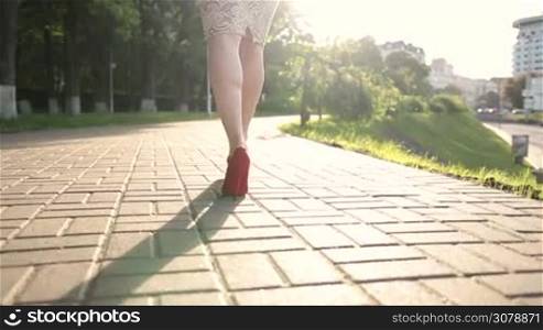 Close-up of elegant slim female legs in classic high heel toe shoes walking along the cobblestone street at sunset. Rear view. Fashionable woman wearing red high heels stepping on the city street. Slow motion. Steadicam stabilized shot.