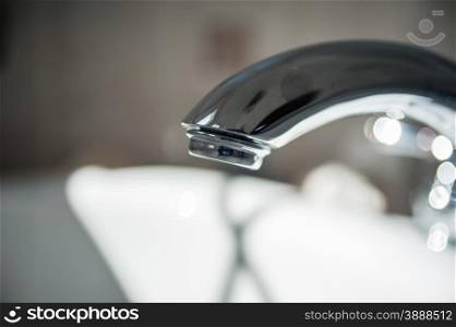 Close-up of end of reflective metallic bathroom faucet against blurred bright and dark background.