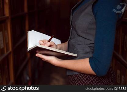 Close up of female hands making notes in notebook with vintage bookshelves on the background.