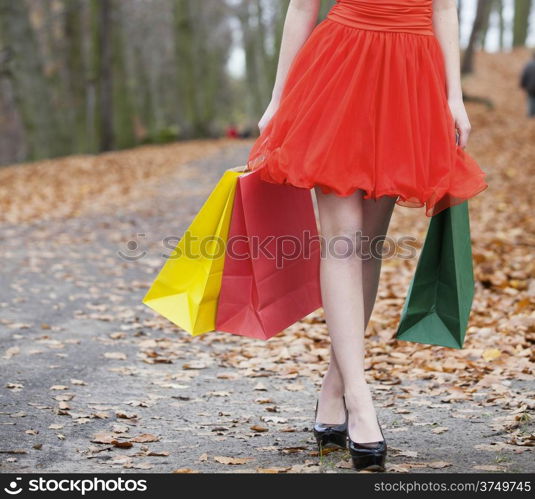 Close up of female legs. Girl in elegant red dress with colorful paper shopping bags walking in the autumnal park. Sale and retail.