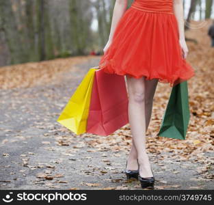 Close up of female legs. Girl in elegant red dress with colorful paper shopping bags walking in the autumnal park. Sale and retail.