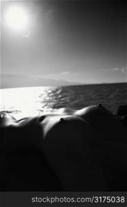 Close up of Filipino young nude woman lying on back against ocean and sun in sky.