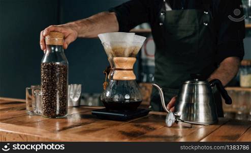 Close up of filter coffee maker, kettle with thermometer and digital scale on wooden table. Barista with tattooed arms wearing dark uniform.