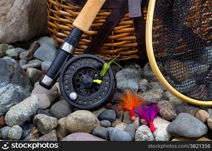 Close up of fishing fly reel, landing net, creel and assorted flies on dry river bed rocks