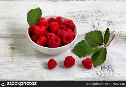 Close up of fresh raspberries, inside and outside of bowl, on rustic white wooden table.