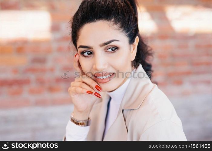 Close up of glamour brunette female having thin eyebrows, dark eyes with long eyelashes, pure skin, full well-shaped lips, red manicure wearing elegant watch on hand and white jacket looking aside
